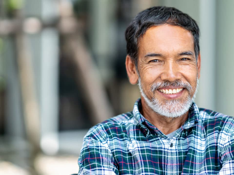 Older man with gray and white beard and plaid shirt smiling outside dental implants washington mi dentist